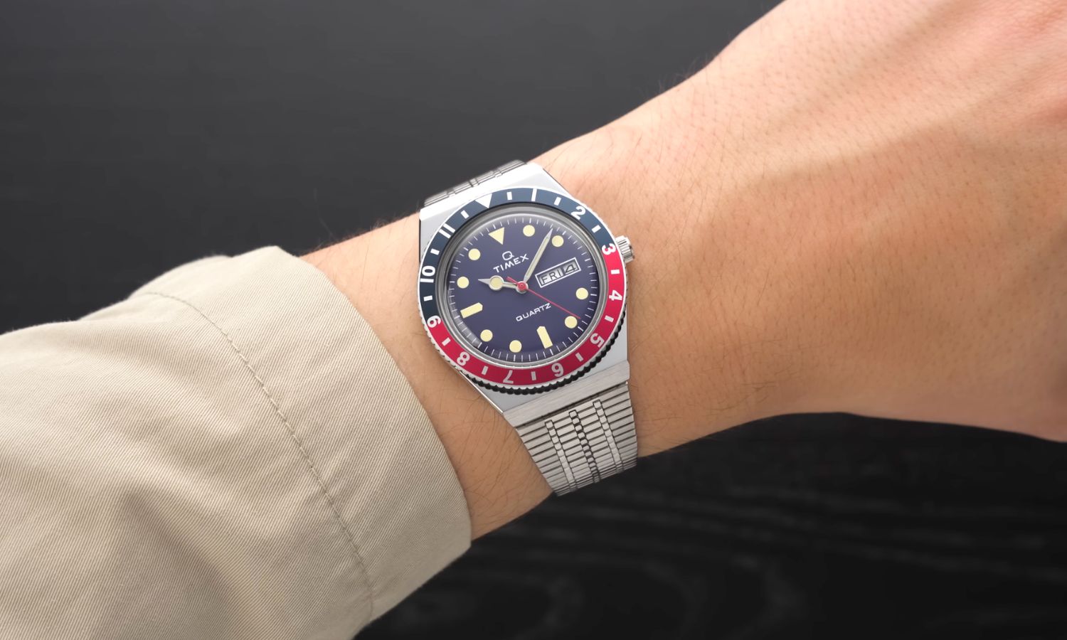 Timex Q Reissue mit dunkelblauem Zifferblatt am Edelstahlarmband an einem Handgelenk mit beigem Ärmel getragen vor schwarzem Hintergrund