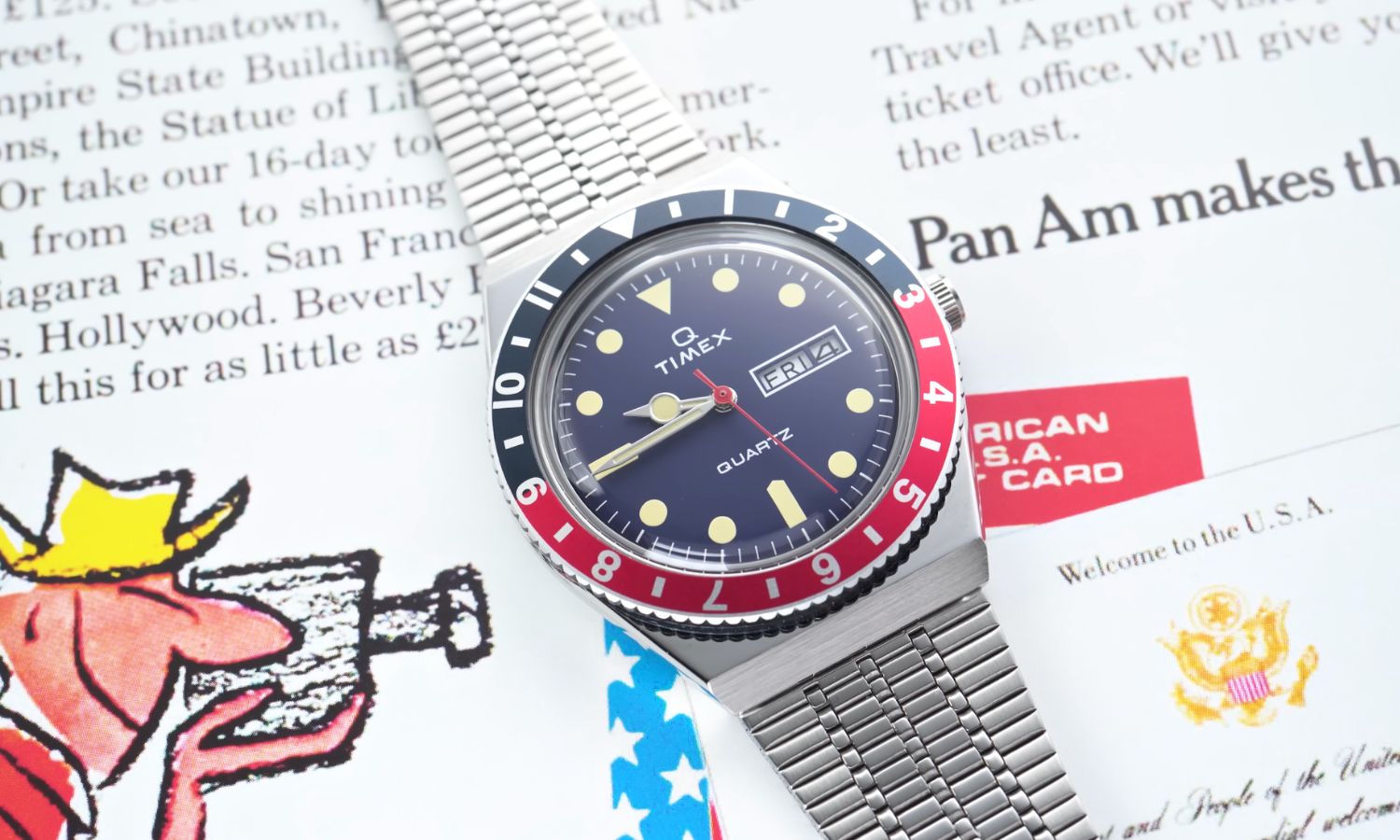 Timex Q Reissue mit dunkelblauem Zifferblatt am Edelstahlarmband auf einer weißen Magazinseite mit schwarzem Text und einer gezeichneten Abbildung eines Kameramanns liegend