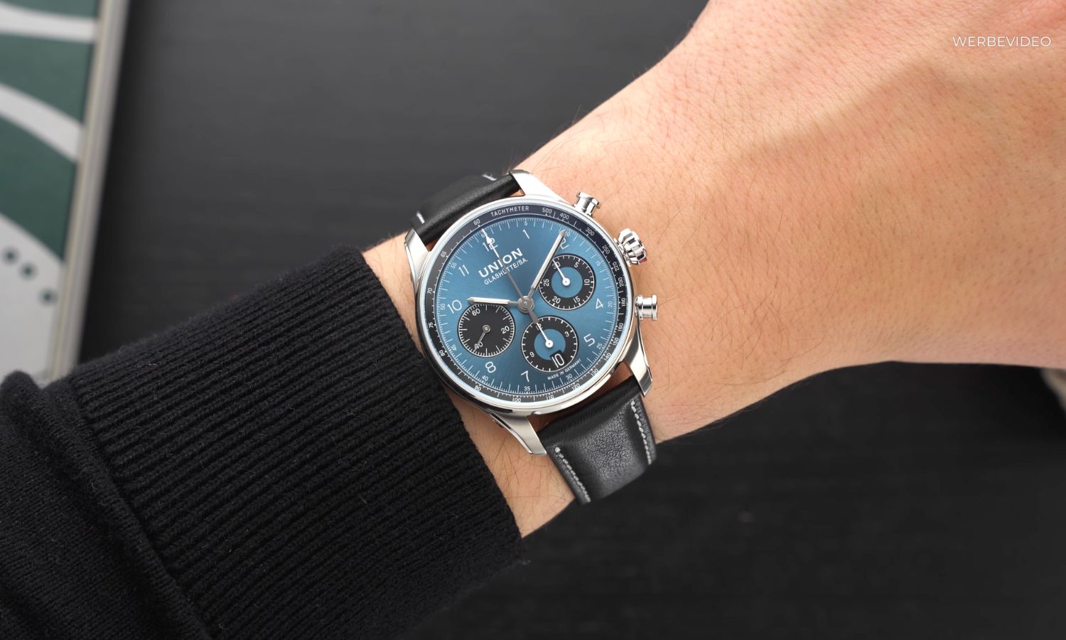 UNION Glashütte Belisar Chronograph mit blauem Zifferblatt am schwarzen Lederarmband an einem Handgelenk mit schwarzem Ärmel getragen vor schwarzem Hintergrund abgebildet