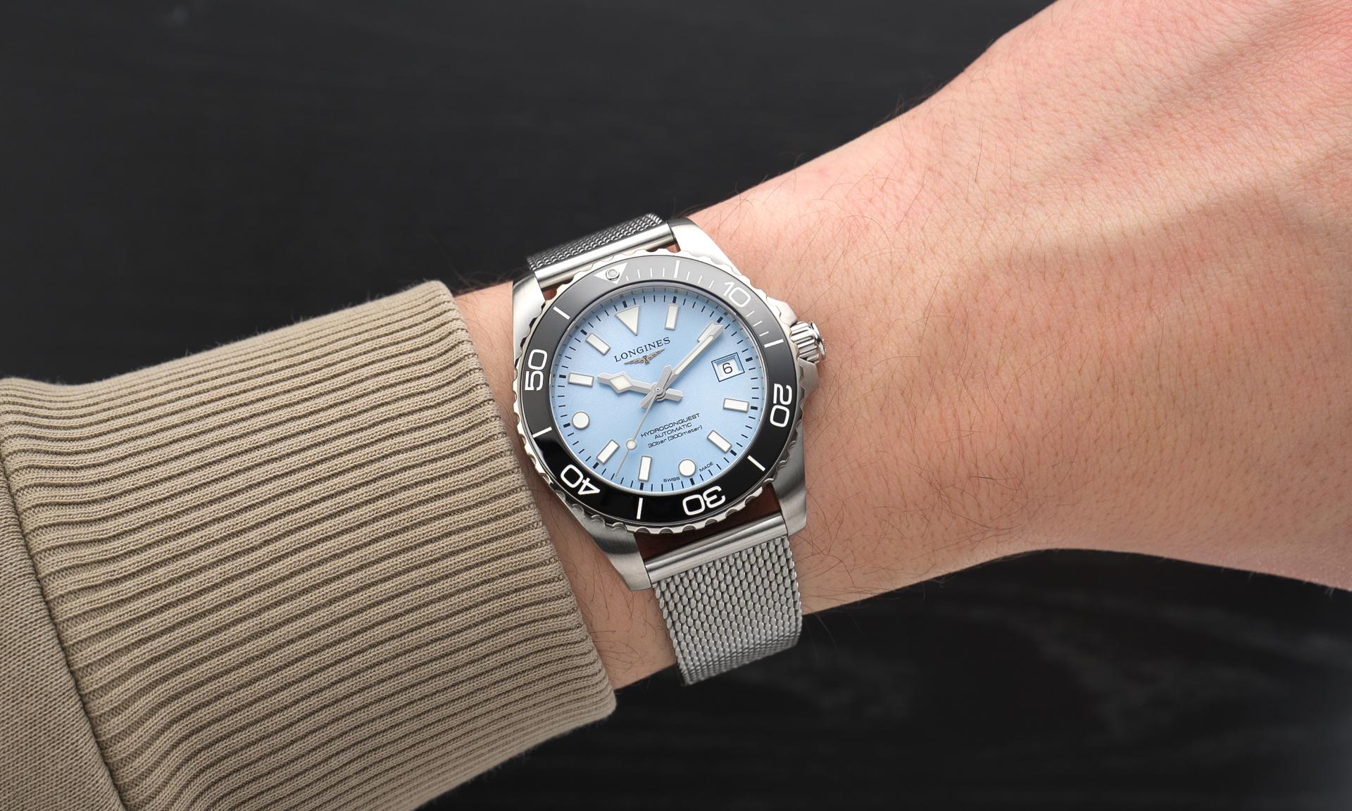 Longines HydroConquest 2026 mit eisblauem Zifferblatt und schwarzer Lünette am Milanaise-Armband an einem Handgelenk mit beigem Ärmel getragen vor schwarzem Hintergrund abgebildet