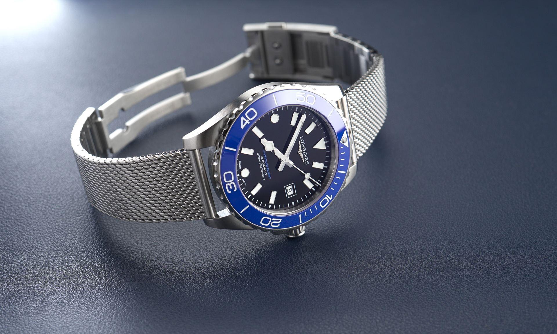 Longines HydroConquest 2026 mit schwarzem Zifferblatt und königsblauer Lünette am Milanaise-Armband auf einem dunkelblauen Untergrund liegend mit geöffneter Schließe