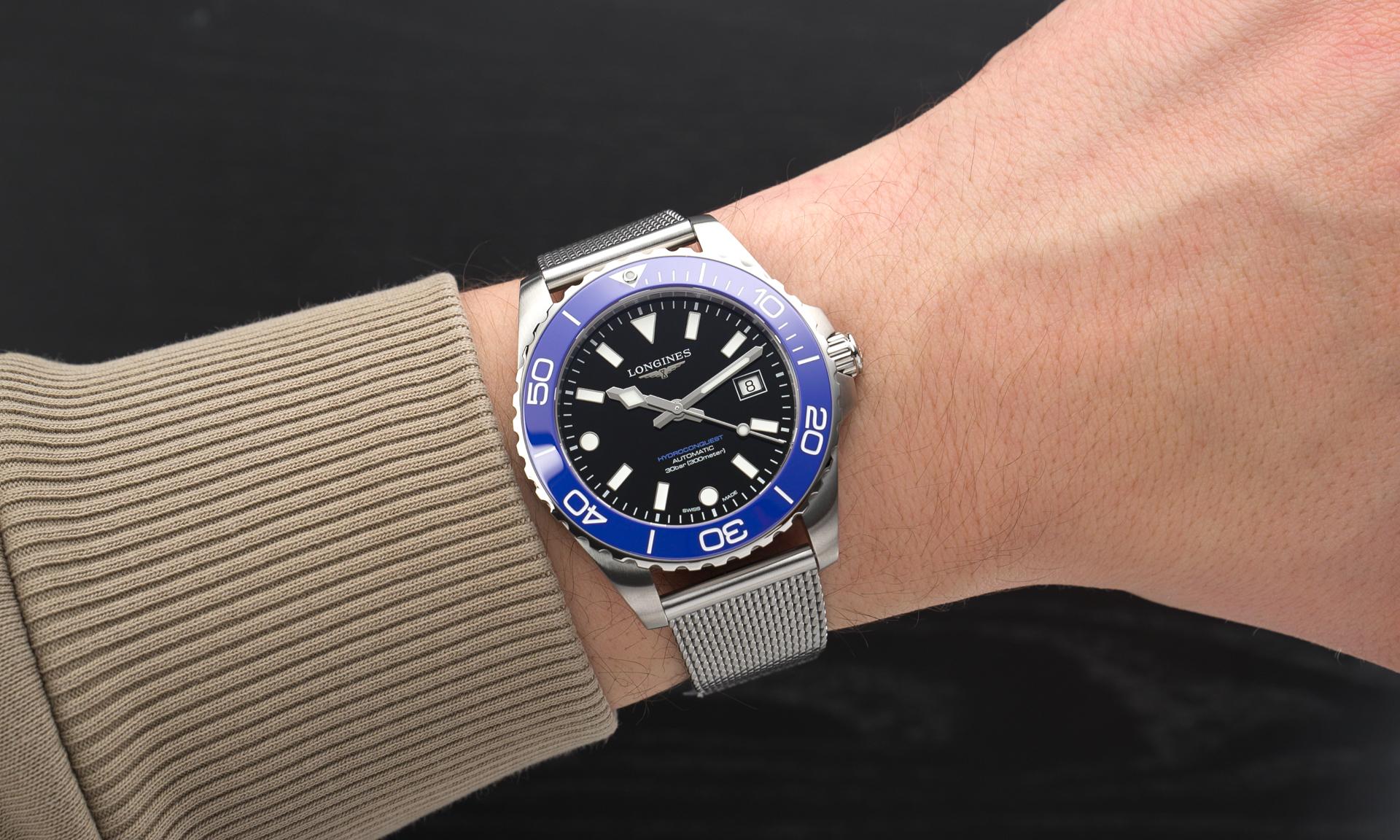 Longines HydroConquest 2026 mit schwarzem Zifferblatt und königsblauer Lünette am Milanaise-Armband an einem Handgelenk mit beigem Ärmel getragen vor schwarzem Hintergrund abgebildet