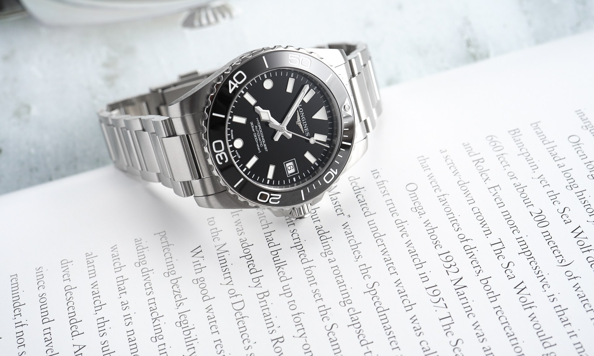 Longines HydroConquest 2026 mit schwarzem Zifferblatt am Edelstahlarmband liegend auf einer weißen Buchseite mit schwarzer Schrift von vorne abgebildet