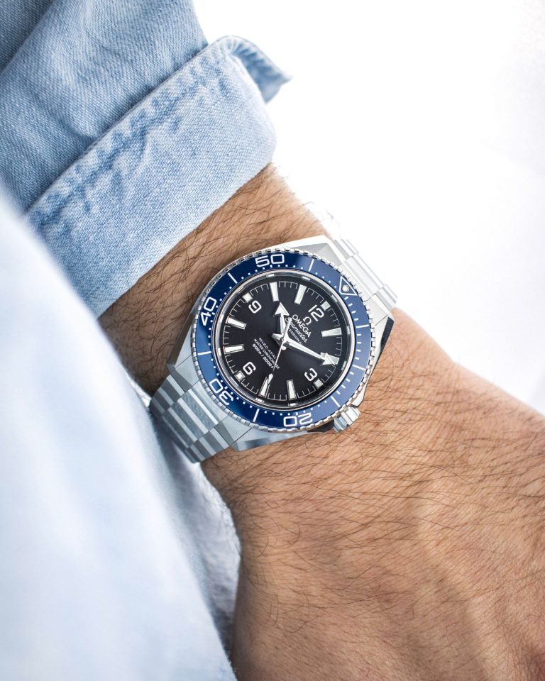 Omega Seamaster Planet Ocean 600M mit schwarzem Zifferblatt und blauer Lünette am Edelstahlarmband an einem Handgelenk mit hellblauem Ärmel getragen vor hellem Hintergrund abgebildet