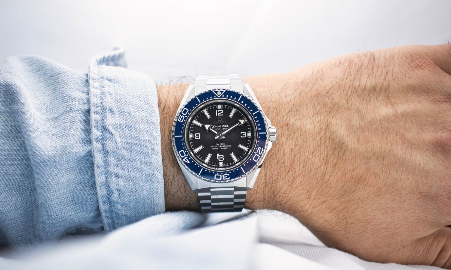 Omega Seamaster Planet Ocean 600M mit schwarzem Zifferblatt und blauer Lünette am Edelstahlarmband an einem Handgelenk mit hellblauem Ärmel getragen vor hellem Hintergrund abgebildet