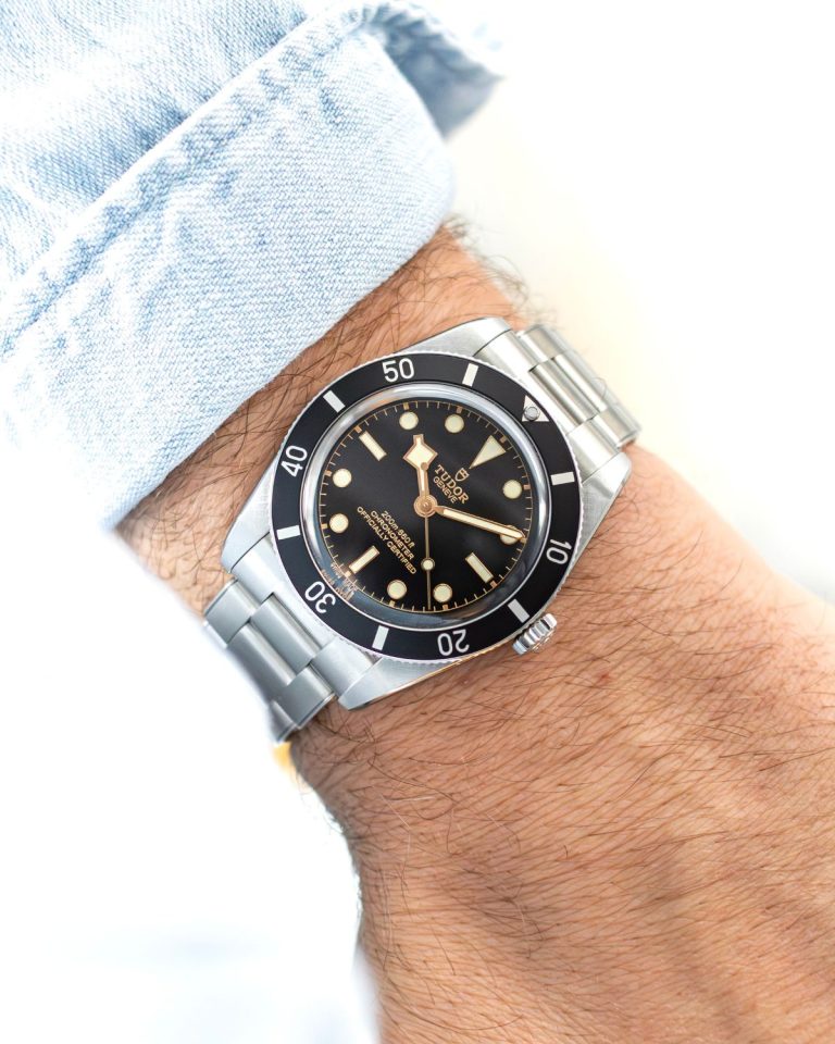 TUDOR Black Bay 54 mit schwarzem Zifferblatt am Edelstahlarmband an einem Handgelenk mit hellblauem Ärmel getragen vor hellem Hintergrund abgebildet
