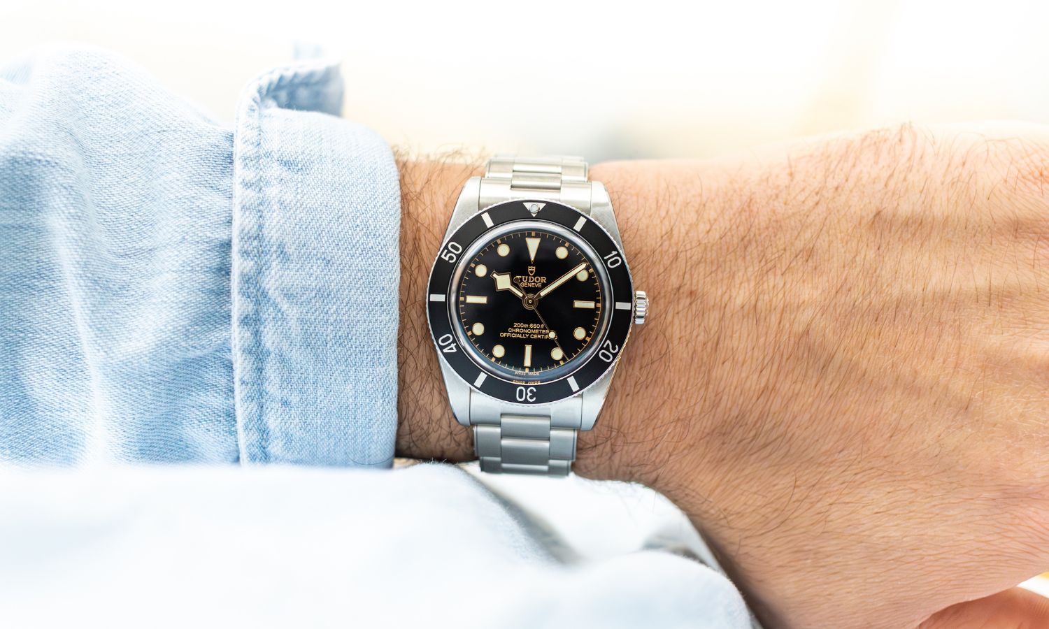TUDOR Black Bay 54 mit schwarzem Zifferblatt am Edelstahlarmband an einem Handgelenk mit hellblauem Ärmel getragen vor hellem Hintergrund abgebildet