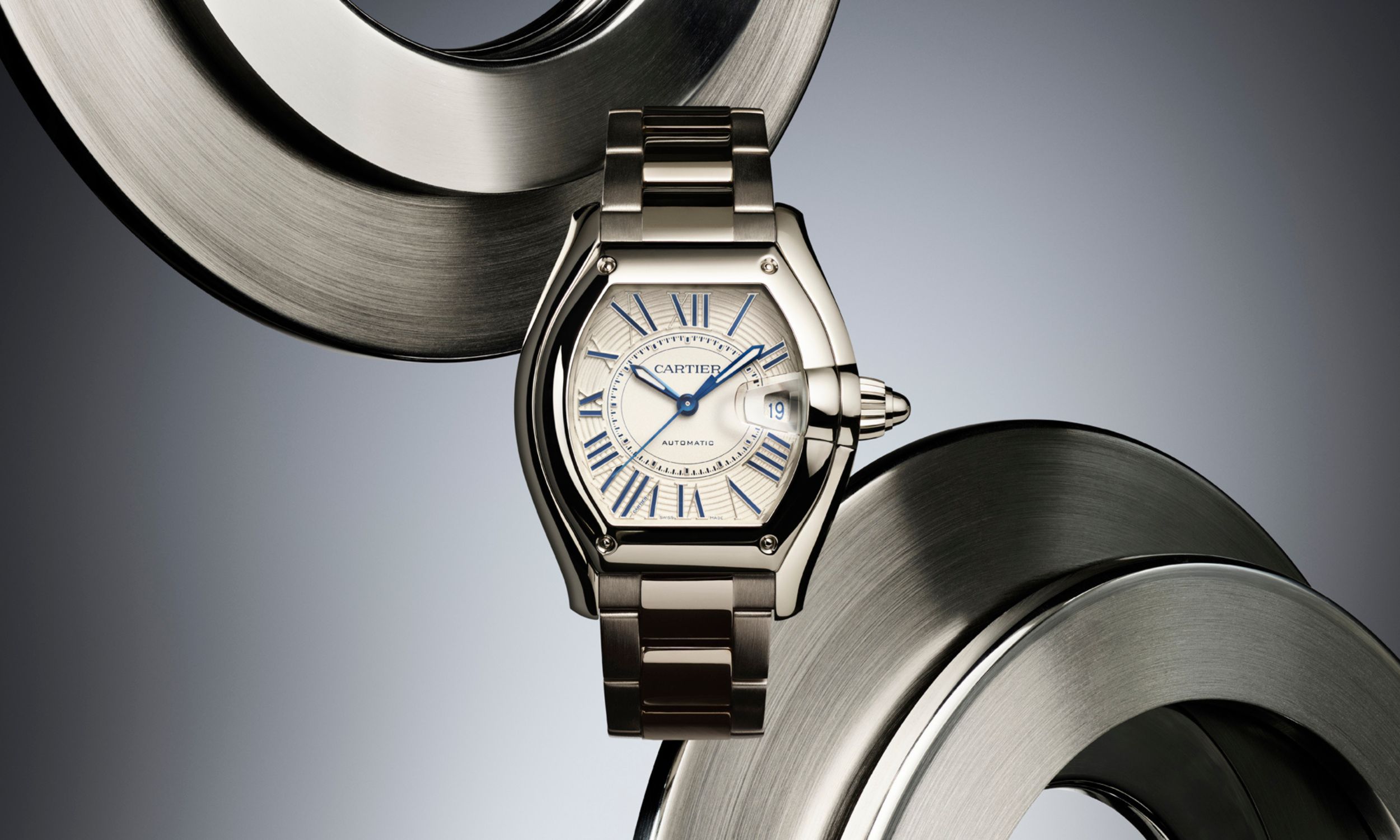 Cartier Roadster vor grauem Hintergrund mit mechanischen Objekten, die wirken wie Bremsscheiben