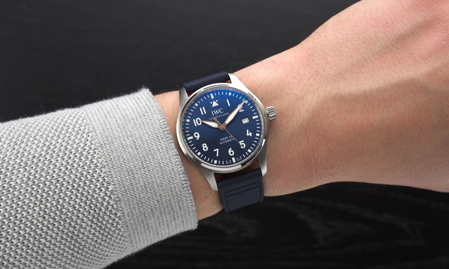 IWC Mark XX Le Petit Prince IW328221 mit dunkelblauem Zifferblatt am dunkelblauen Gummiband an einem Handgelenk mit grauem Ärmel getragen vor schwarzem Hintergrund