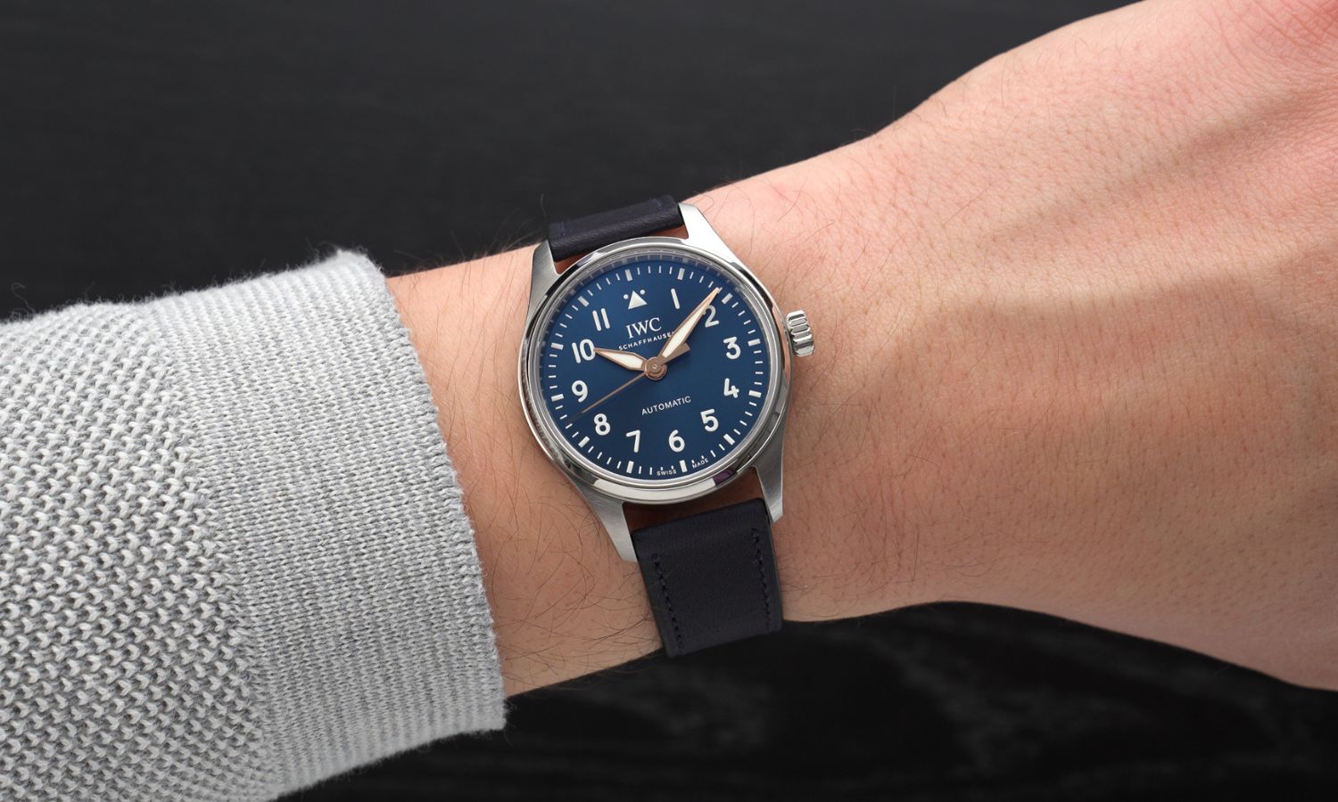 IWC Pilot's Watch 36 Le Petit Prince IW458802 mit dunkelblauem Zifferblatt am dunkelblauen Gummiband an einem Handgelenk mit grauem Ärmel getragen vor schwarzem Hintergrund