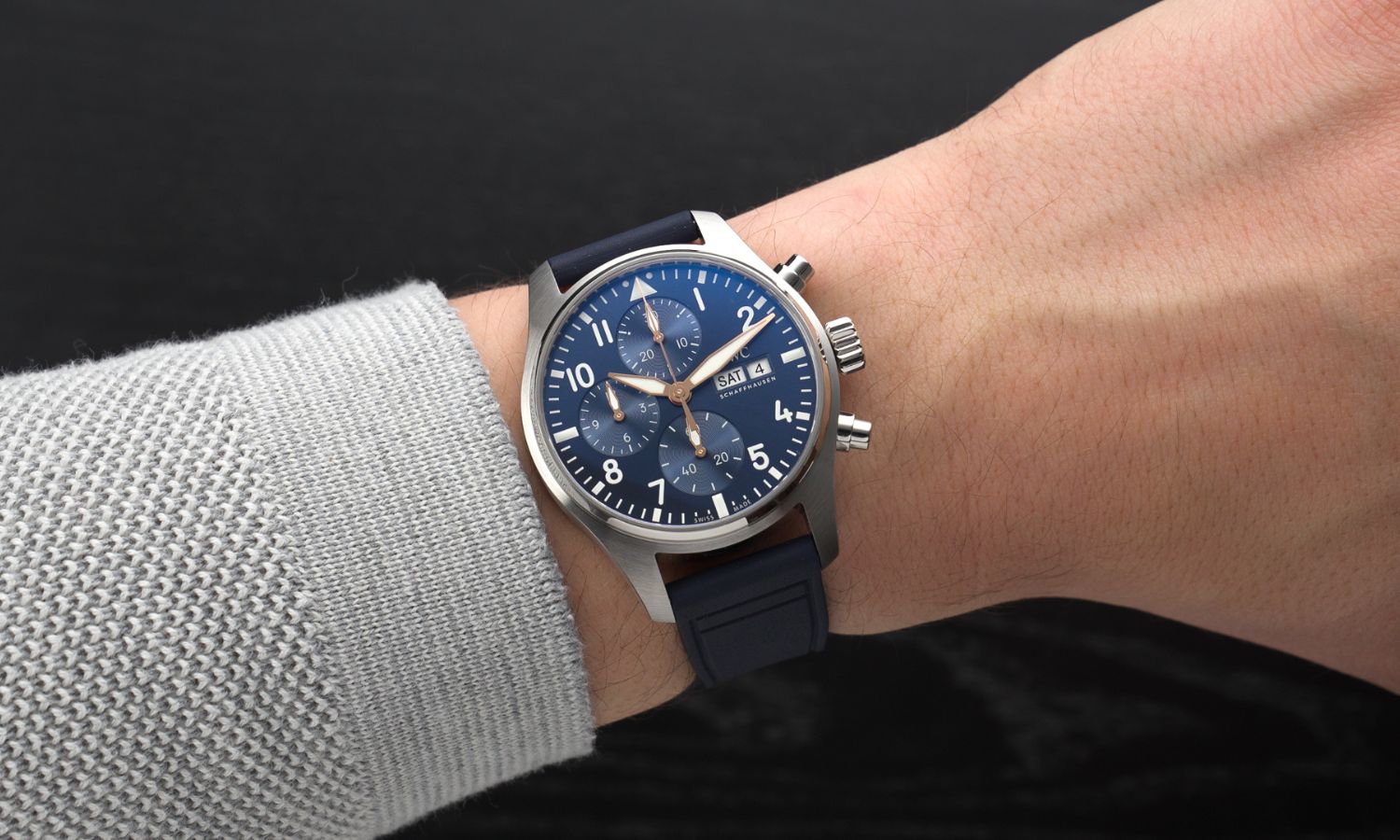 IWC Pilot's Watch Chronograph 41 Le Petit Prince IW388120 mit dunkelblauem Zifferblatt am dunkelblauen Gummiband an einem Handgelenk mit grauem Ärmel getragen vor schwarzem Hintergrund