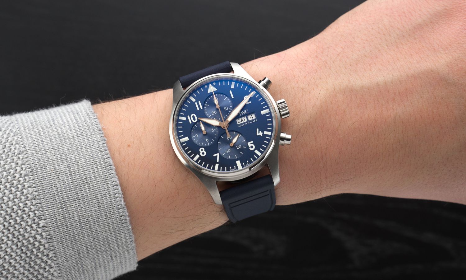 IWC Pilot's Watch Chronograph 43 Le Petit Prince IW378011 mit dunkelblauem Zifferblatt am dunkelblauen Gummiband an einem Handgelenk mit grauem Ärmel getragen vor schwarzem Hintergrund