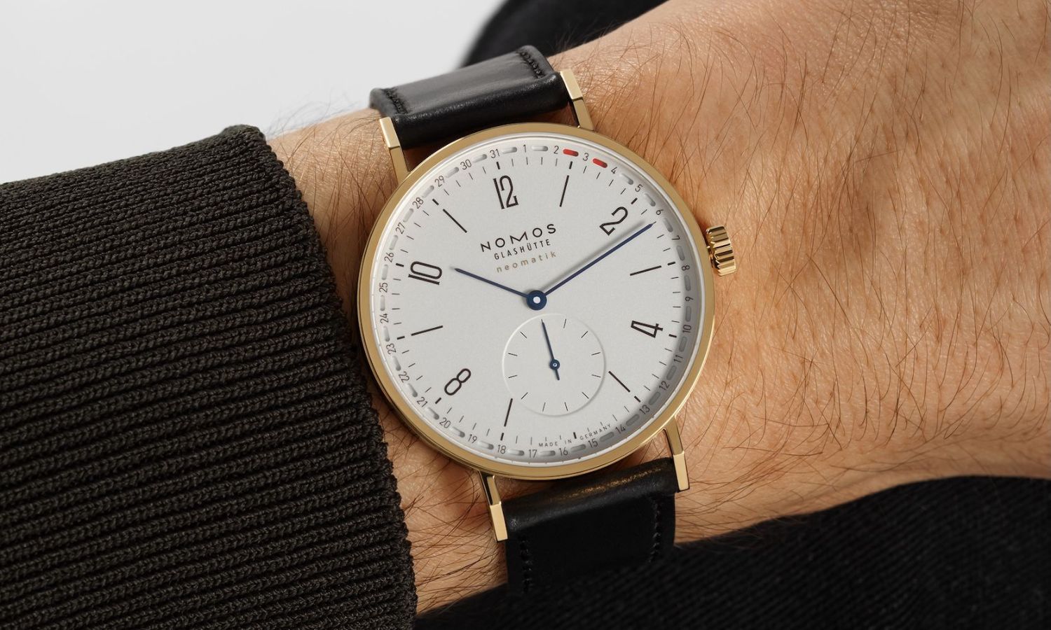 NOMOS Glashütte Tangente neomatik 38 Update Gold mit weißem Zifferblatt am schwarzen Lederarmband an einem Handgelenk mit schwarzem Ärmel getragen