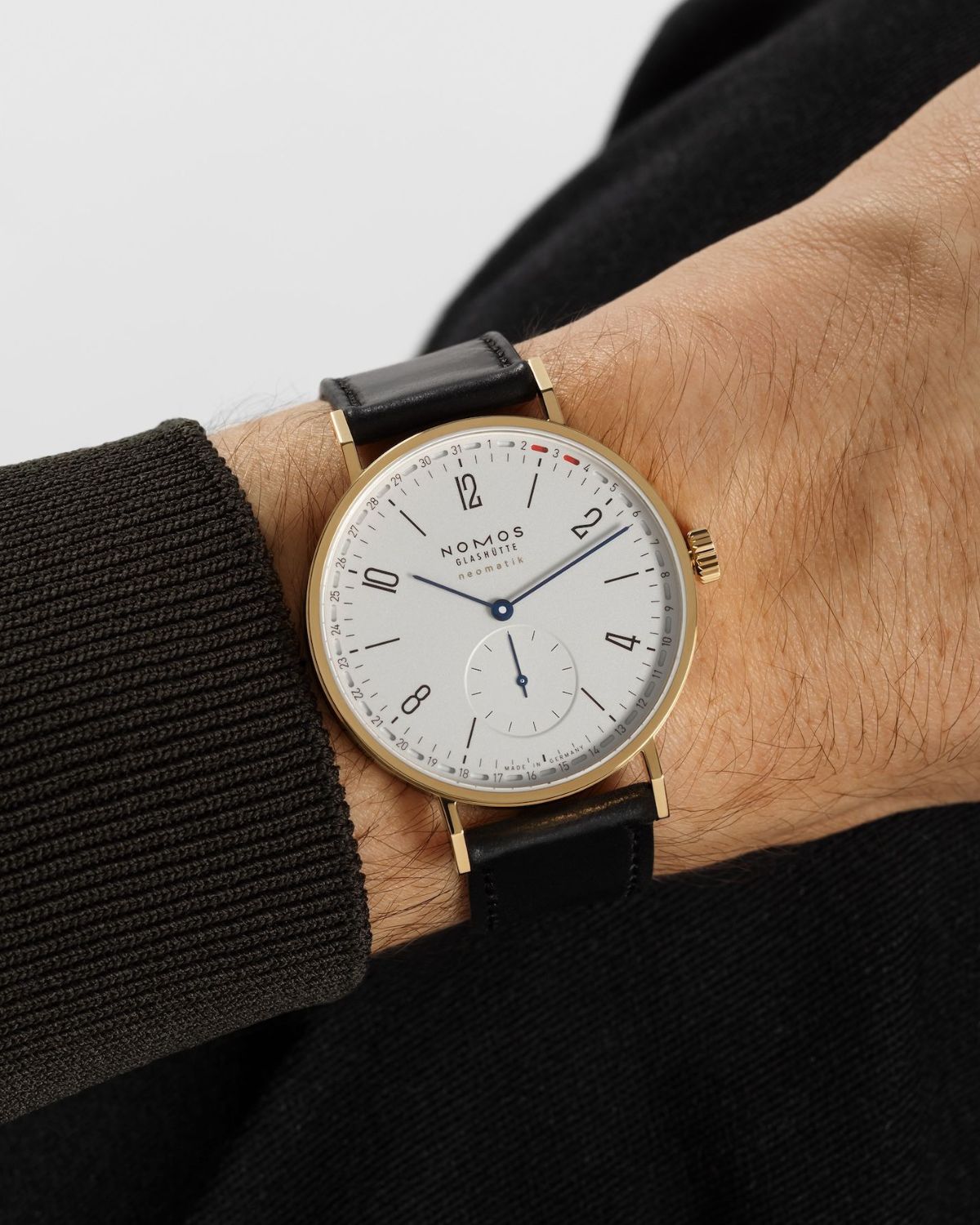 NOMOS Glashütte Tangente neomatik 38 Update Gold mit weißem Zifferblatt am schwarzen Lederarmband an einem Handgelenk mit schwarzem Ärmel getragen
