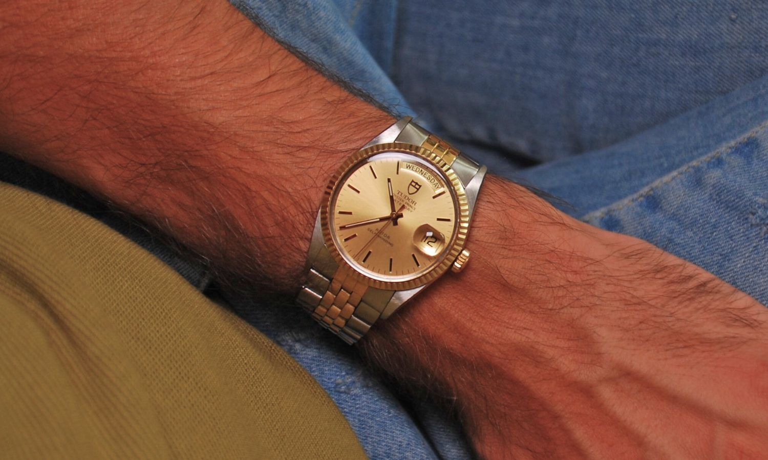 TUDOR Oyster Prince Date-Day Referenz 94613 mit goldenem Zifferblatt am Edelstahlarmband an einem Handgelenk getragen mit einer Jeanshose im Hintergrund
